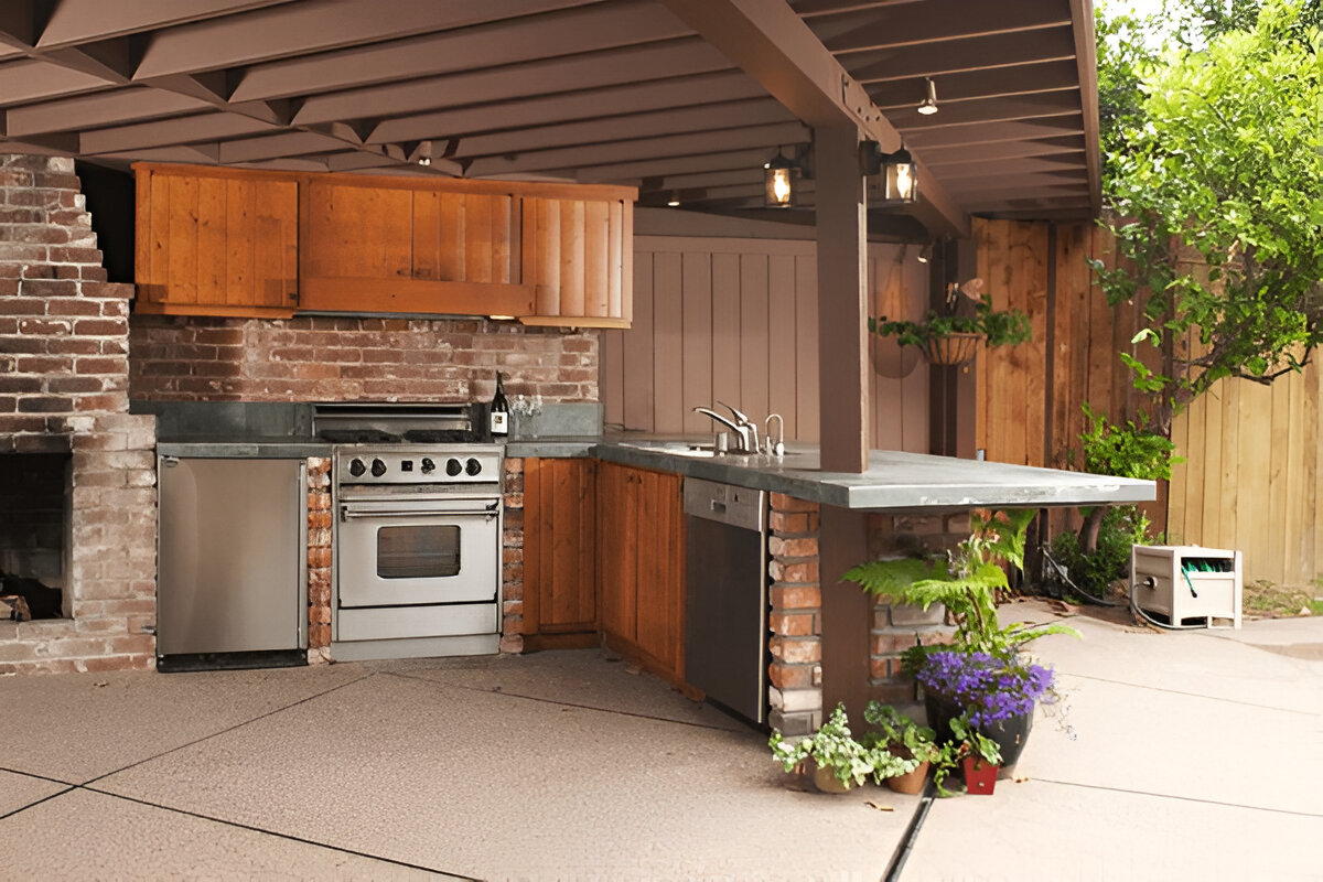 Rustic outdoor kitchen with exposed brick walls, wooden cabinets, a concrete countertop, stainless steel appliances, and a pergola with hanging lanterns, surrounded by lush greenery