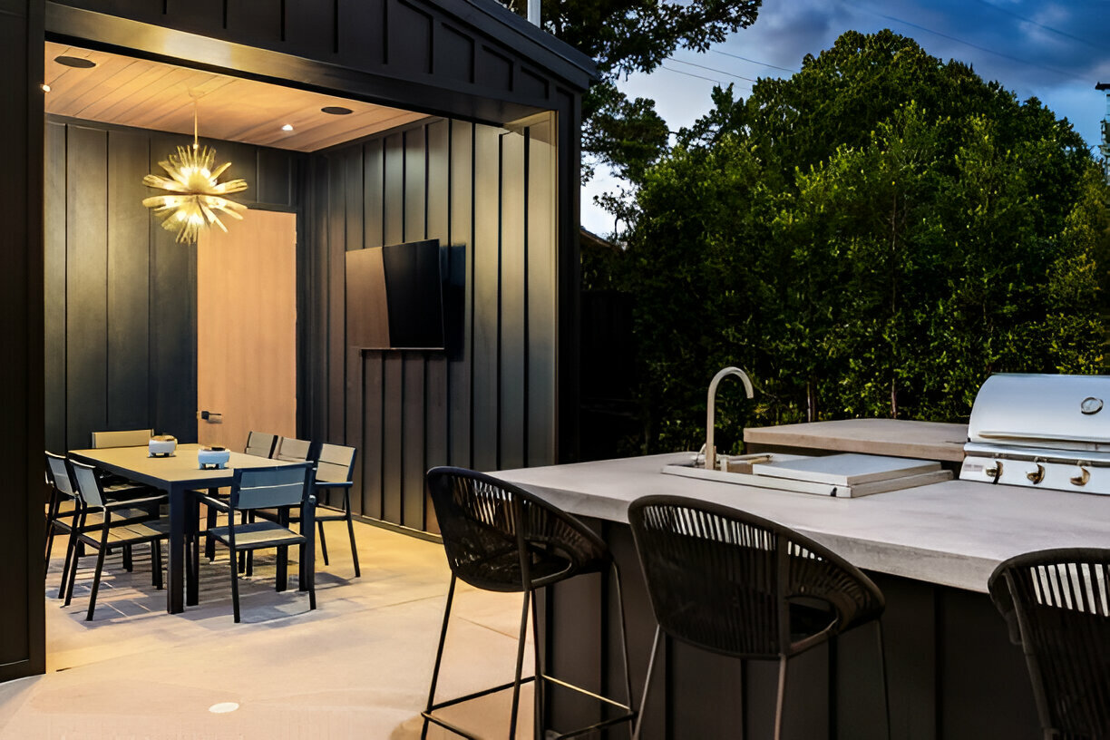 Modern outdoor kitchen with a black bar, stainless steel grill, and sink, complemented by a dining area with a gold chandelier and outdoor TV. Surrounded by lush greenery for a cozy ambiance