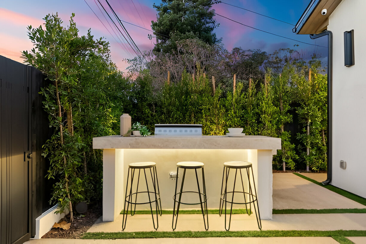 Minimalist outdoor bar with a concrete countertop, ambient under-counter lighting, and three black metal stools, set against lush greenery at twilight