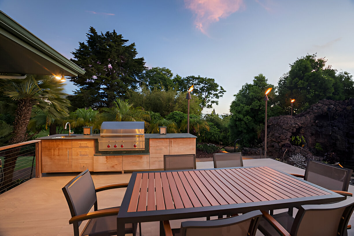 Luxury outdoor kitchen with a built-in stainless steel grill, sleek cabinetry, and a spacious wood-accented dining table, set against a tropical backdrop with tiki torches