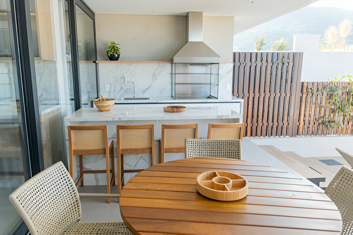 Modern outdoor kitchen with marble countertops, a stainless steel range hood, wooden bar stools, and a cozy dining area with woven chairs and a round wooden table