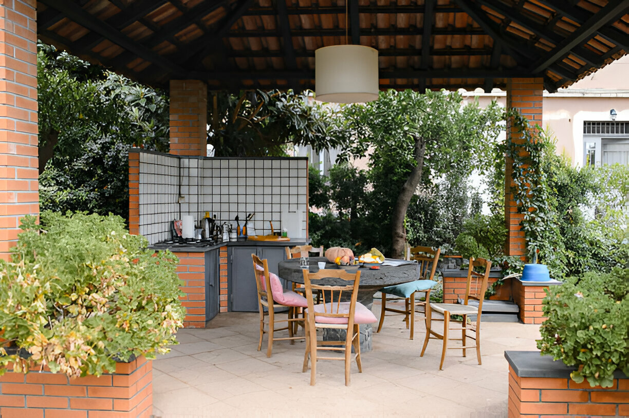 Rustic outdoor kitchen with a tiled counter, wooden dining table, and mismatched chairs, surrounded by greenery under a terracotta roof