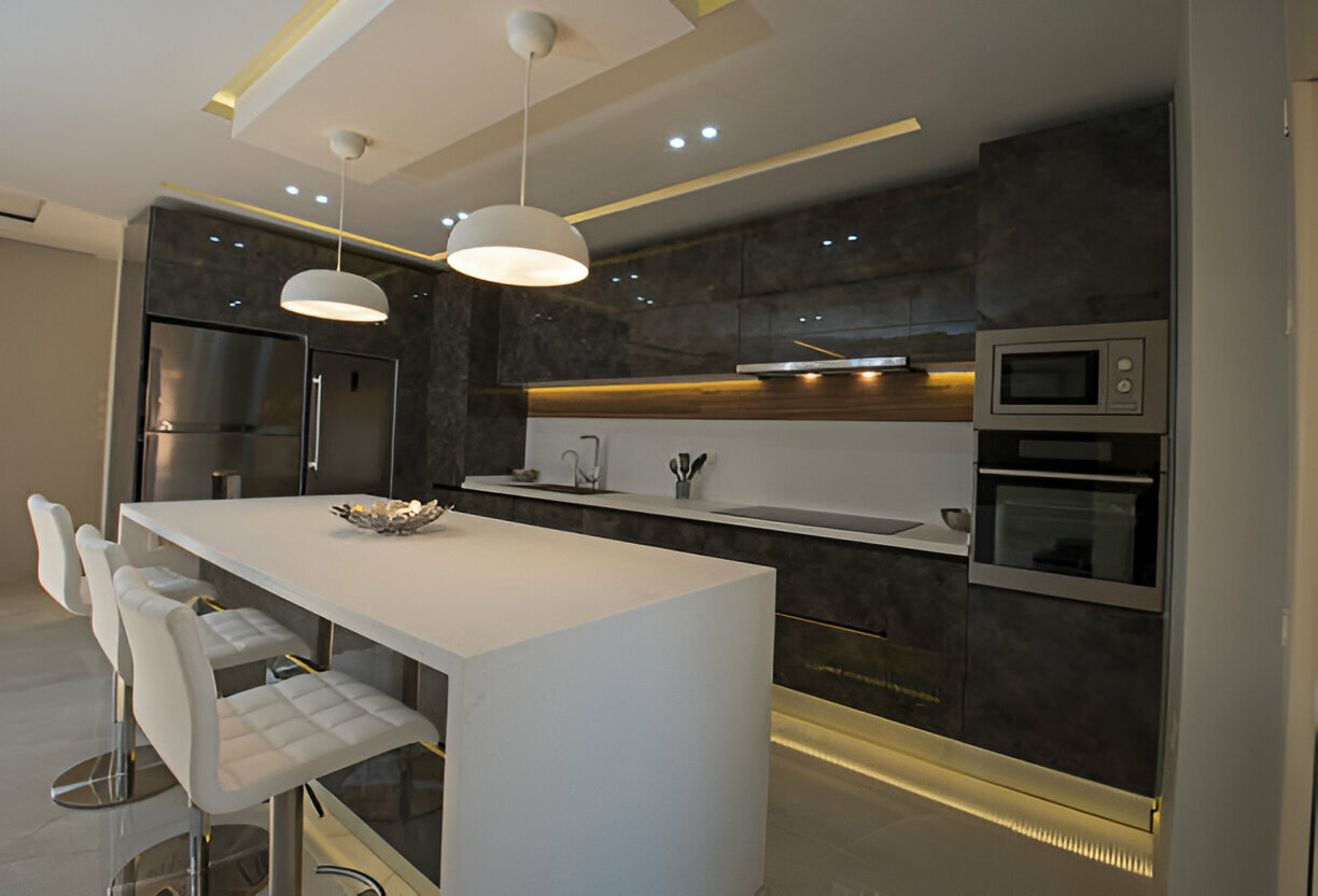 Modern luxury kitchen with dark marble cabinets, white waterfall island, pendant lighting, and built-in stainless steel appliances. LED under-cabinet lighting enhances the ambiance
