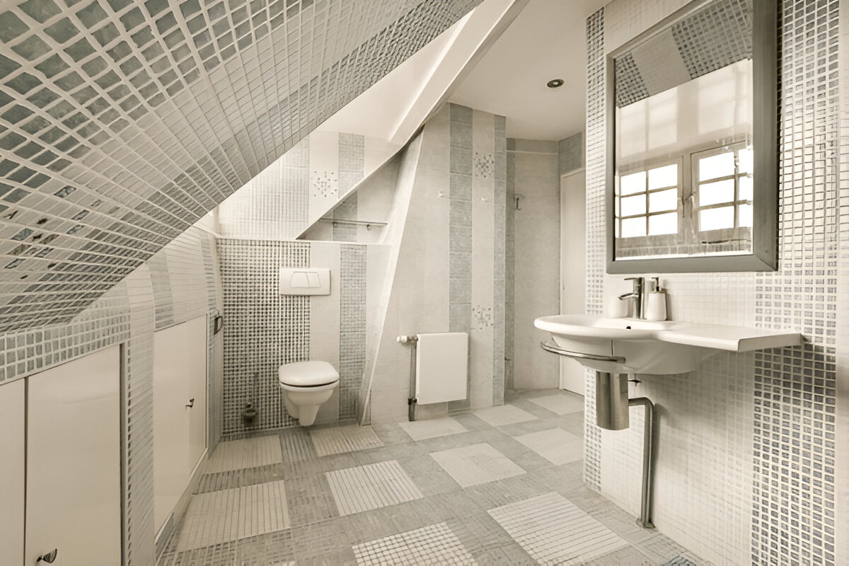 A modern attic bathroom with mosaic and patterned tiles, a wall-mounted toilet, and a floating sink under a slanted ceiling.