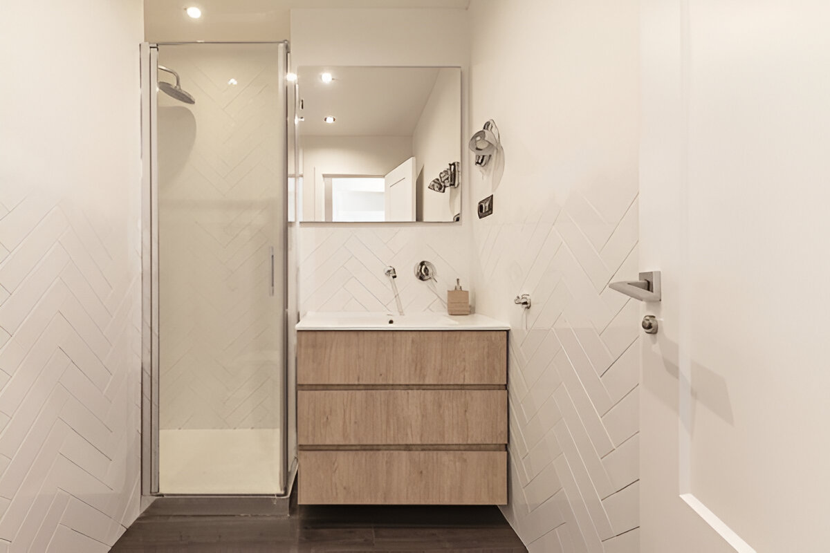 Compact bathroom featuring a wood vanity, white herringbone tiles, and a glass-enclosed shower