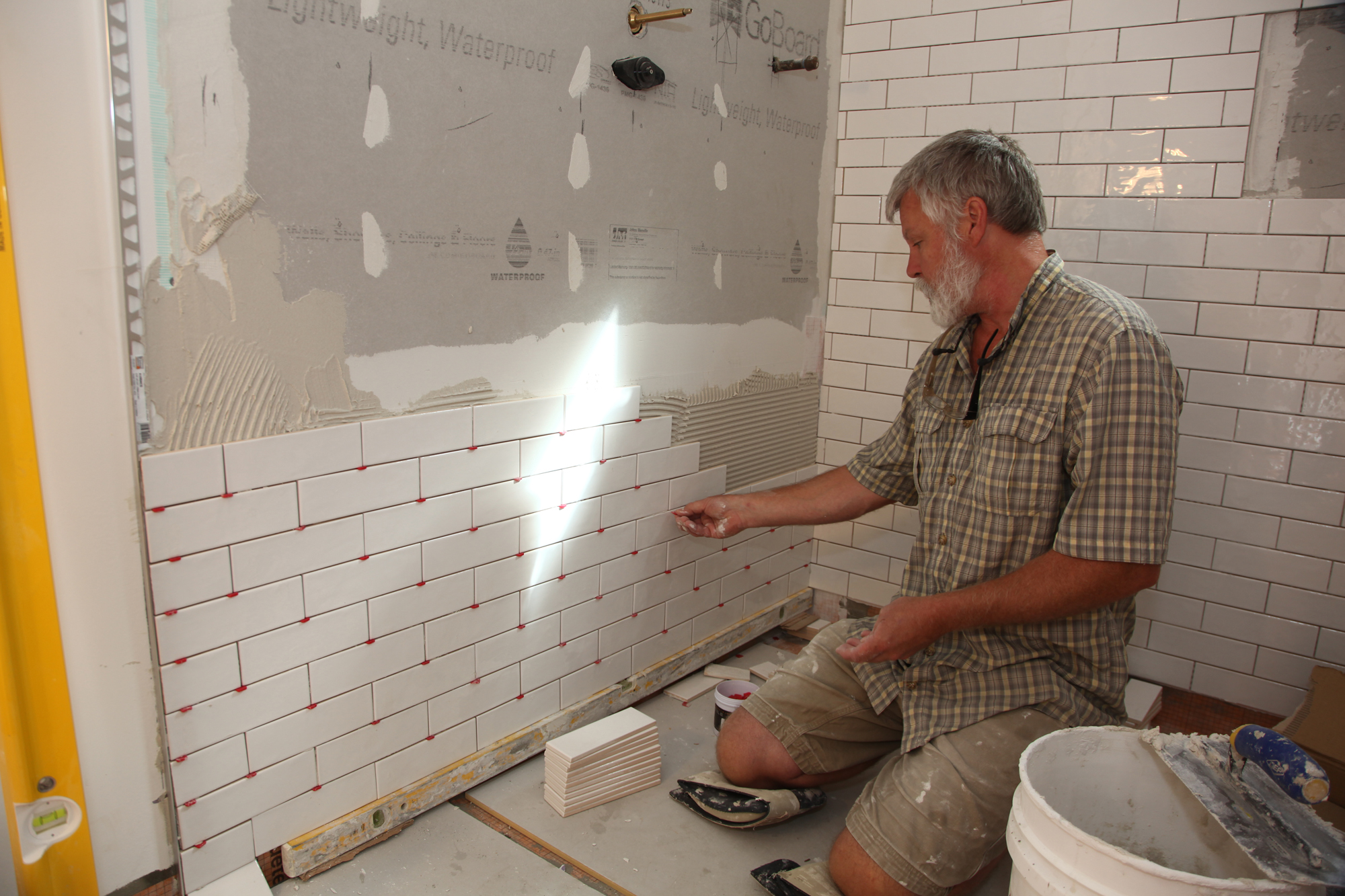 The image shows an elderly man working on tiling the walls of a bathroom. The walls are partially covered with white subway tiles, and there are construction materials and tools scattered around the room