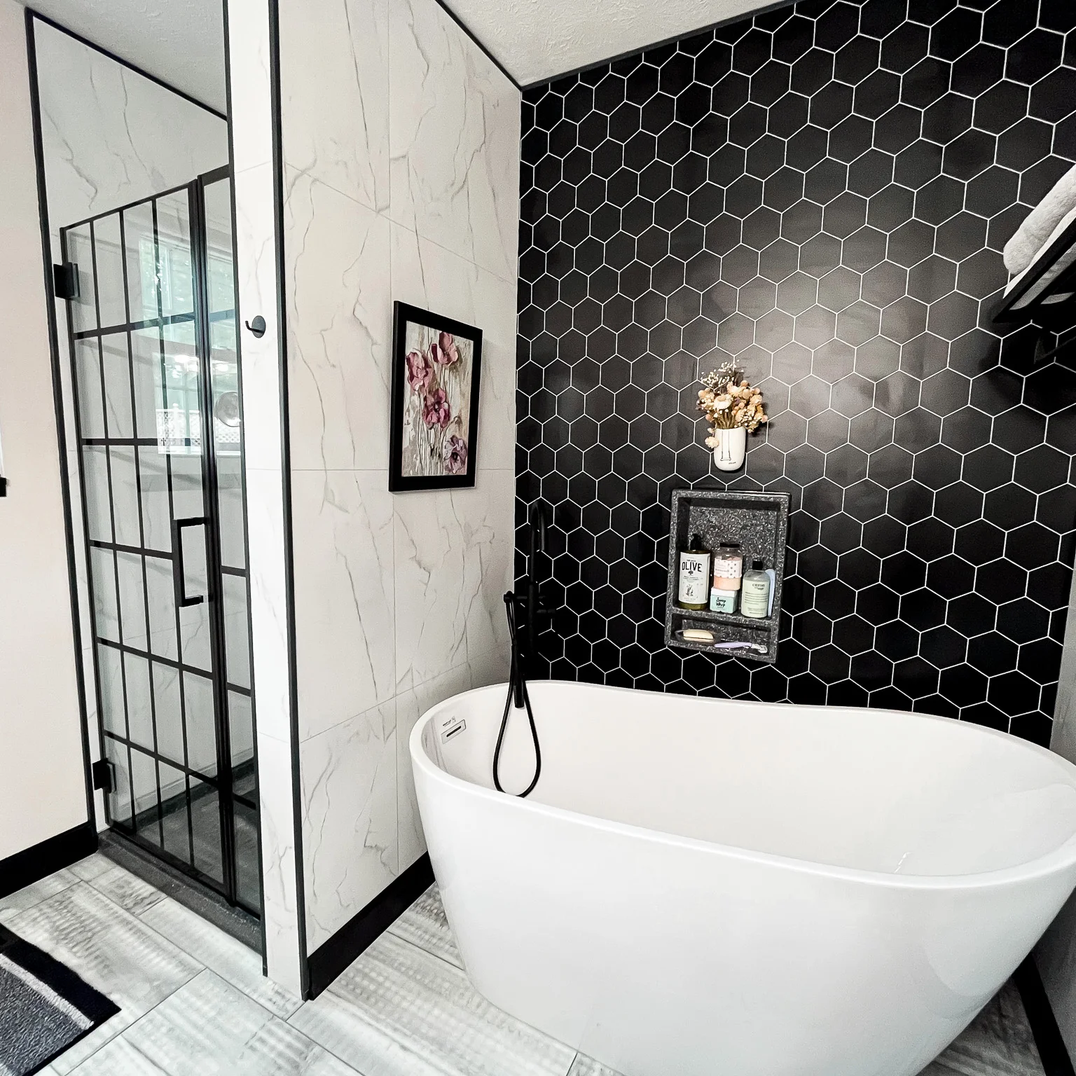 The image shows a modern bathroom with a white freestanding bathtub, a black hexagonal tile wall, and a glass shower enclosure with a black metal frame.