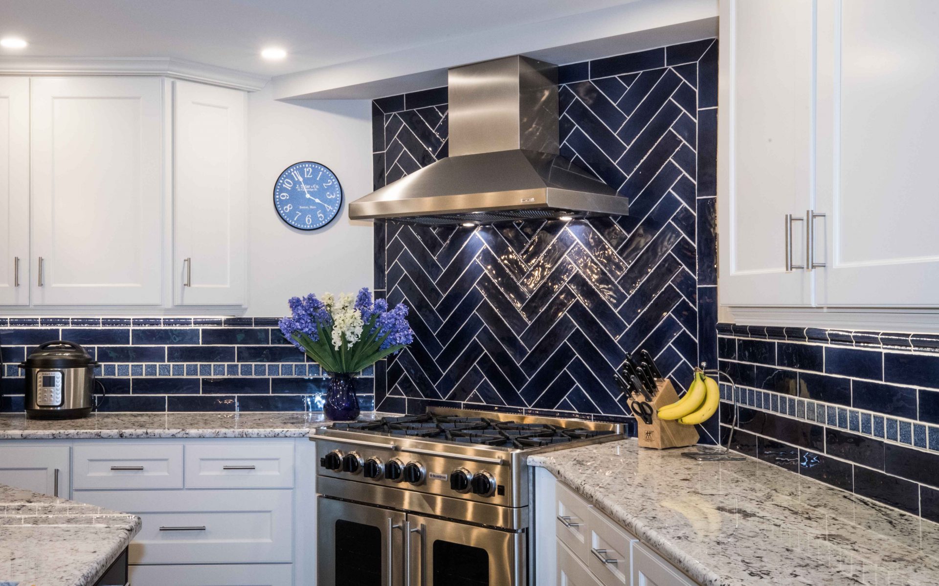 A modern kitchen with a bold, herringbone-patterned backsplash in navy blue tiles, a stainless steel range hood, and white cabinetry. The room features a large central island with a granite countertop
