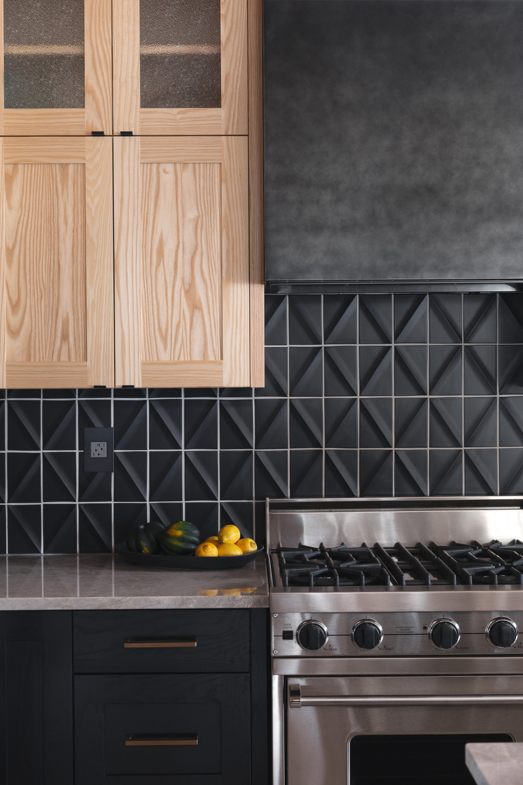 A contemporary kitchen with a sleek and minimalist aesthetic. The image shows a kitchen with dark gray tiles forming a geometric pattern on the backsplash, contrasting with the natural wood cabinetry and drawers. A stainless steel gas range is visible, along with a bowl of fresh produce on the countertop