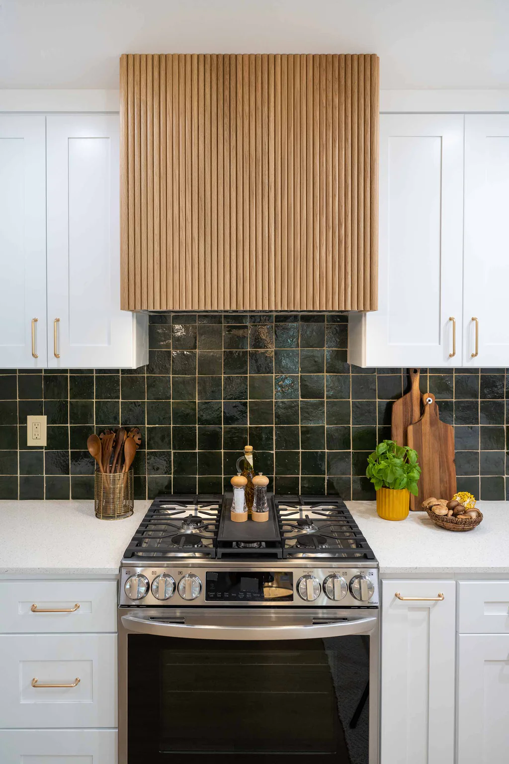 The image shows a modern kitchen with a striking wooden backsplash and a tiled countertop. The wooden backsplash is made up of vertical wooden slats that extend from the countertop to the ceiling, creating a warm and textured backdrop. The countertop features a dark, mottled tile pattern that complements the wood. Various kitchen accessories and utensils are visible on the countertop, adding to the functional and lived-in feel of the space