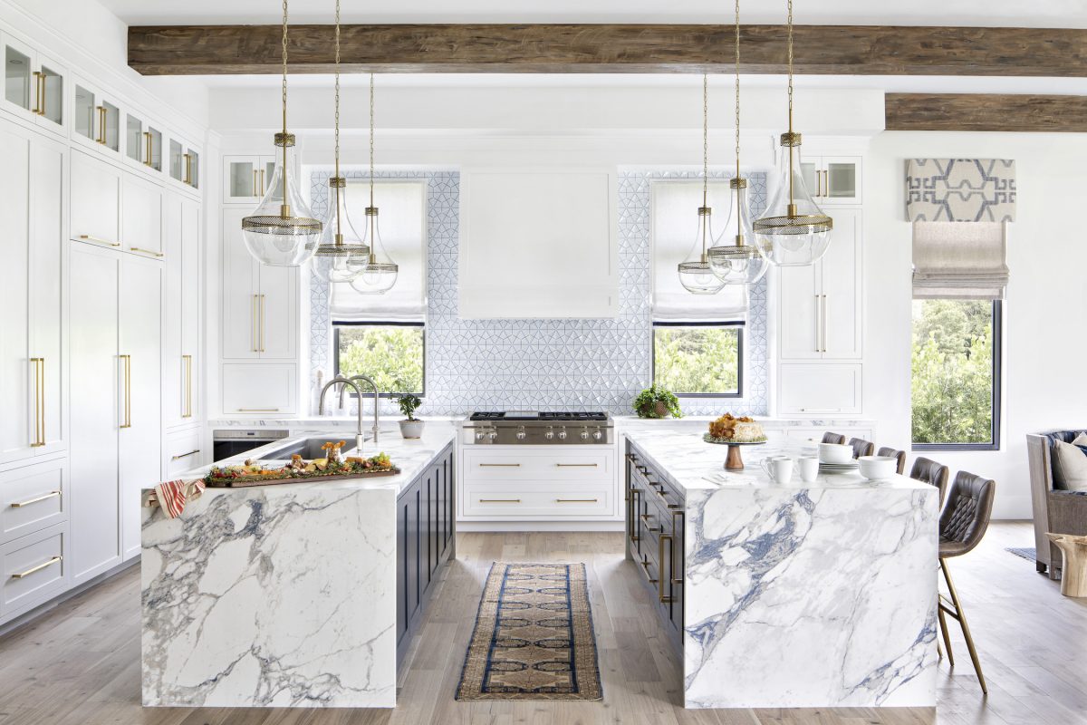 Luxury kitchen featuring dual Arabescato marble waterfall islands, white cabinetry, brass hardware, and exposed ceiling beams