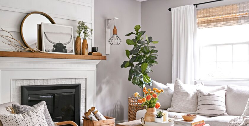 A cozy living room with a white fireplace, neutral-toned furniture, a wooden coffee table with flowers, a leather pouf, and natural light from a window with white curtains and bamboo blinds.