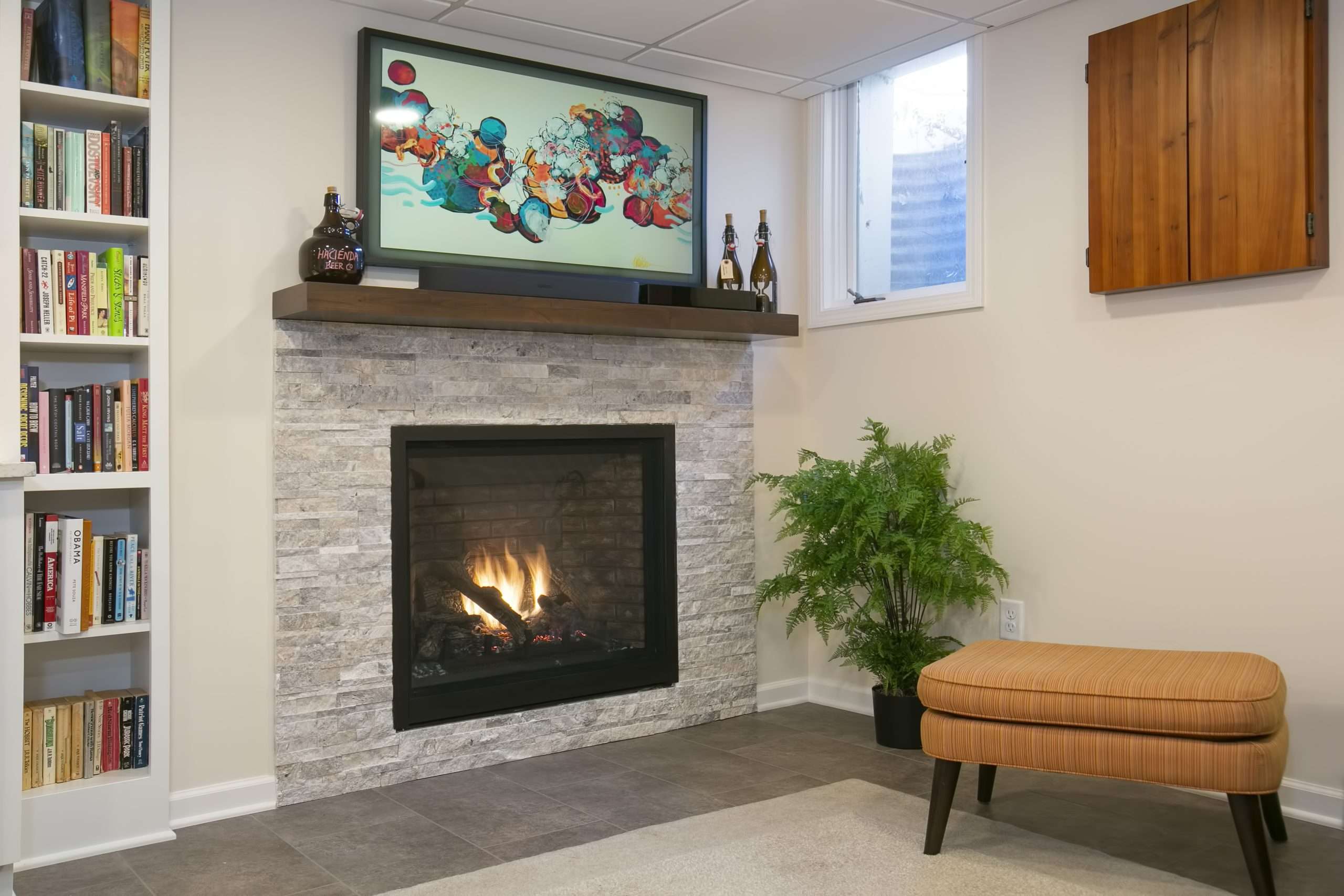 A modern living room with a stone-clad fireplace, a TV and decorative items on the mantle, a bookshelf on the side, and a potted plant adding warmth to the space.
