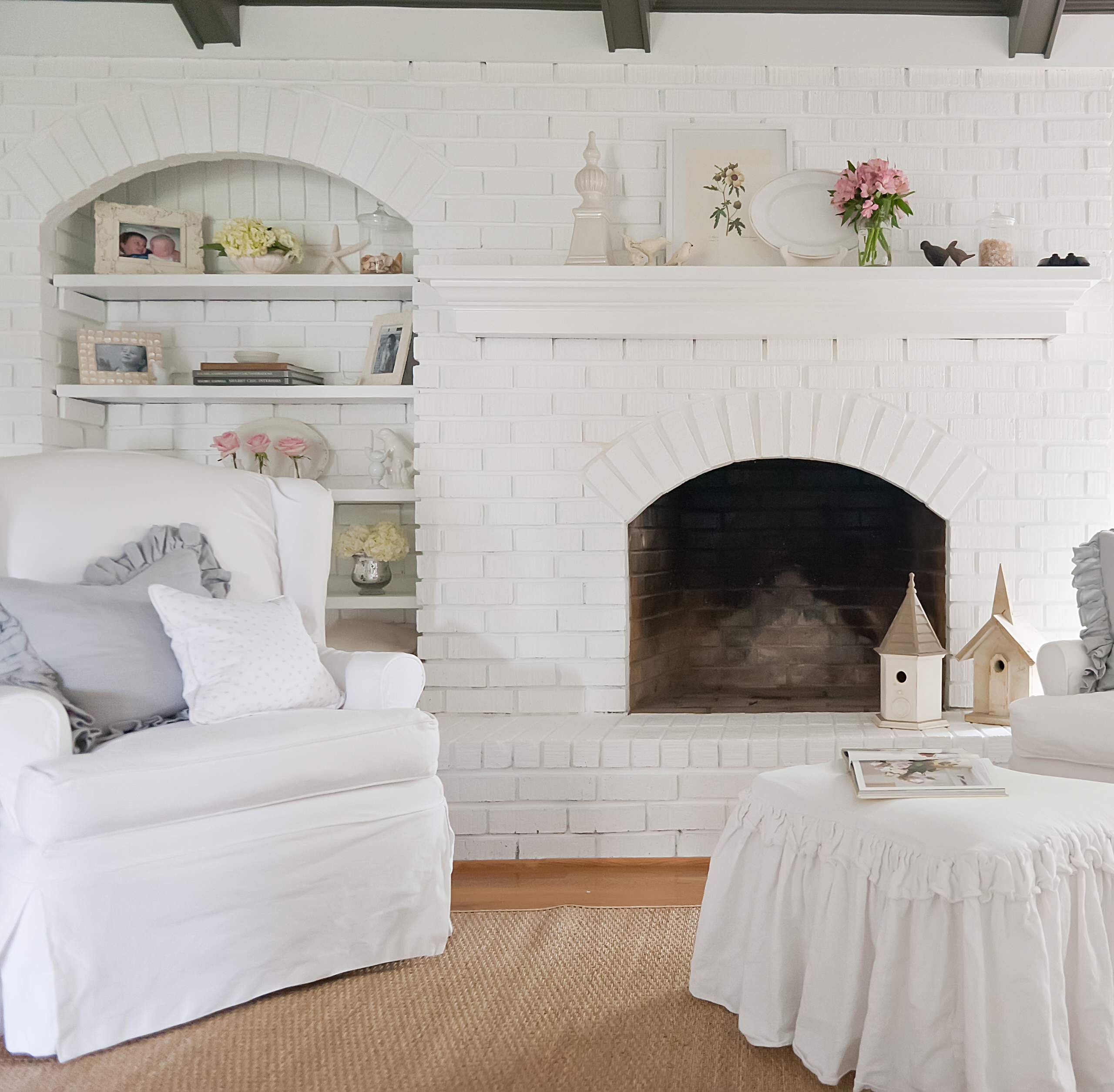 A cozy living room with a white-painted brick fireplace, built-in shelves decorated with flowers, books, and frames, white slipcovered armchairs, a round ottoman, and rustic wooden birdhouses.