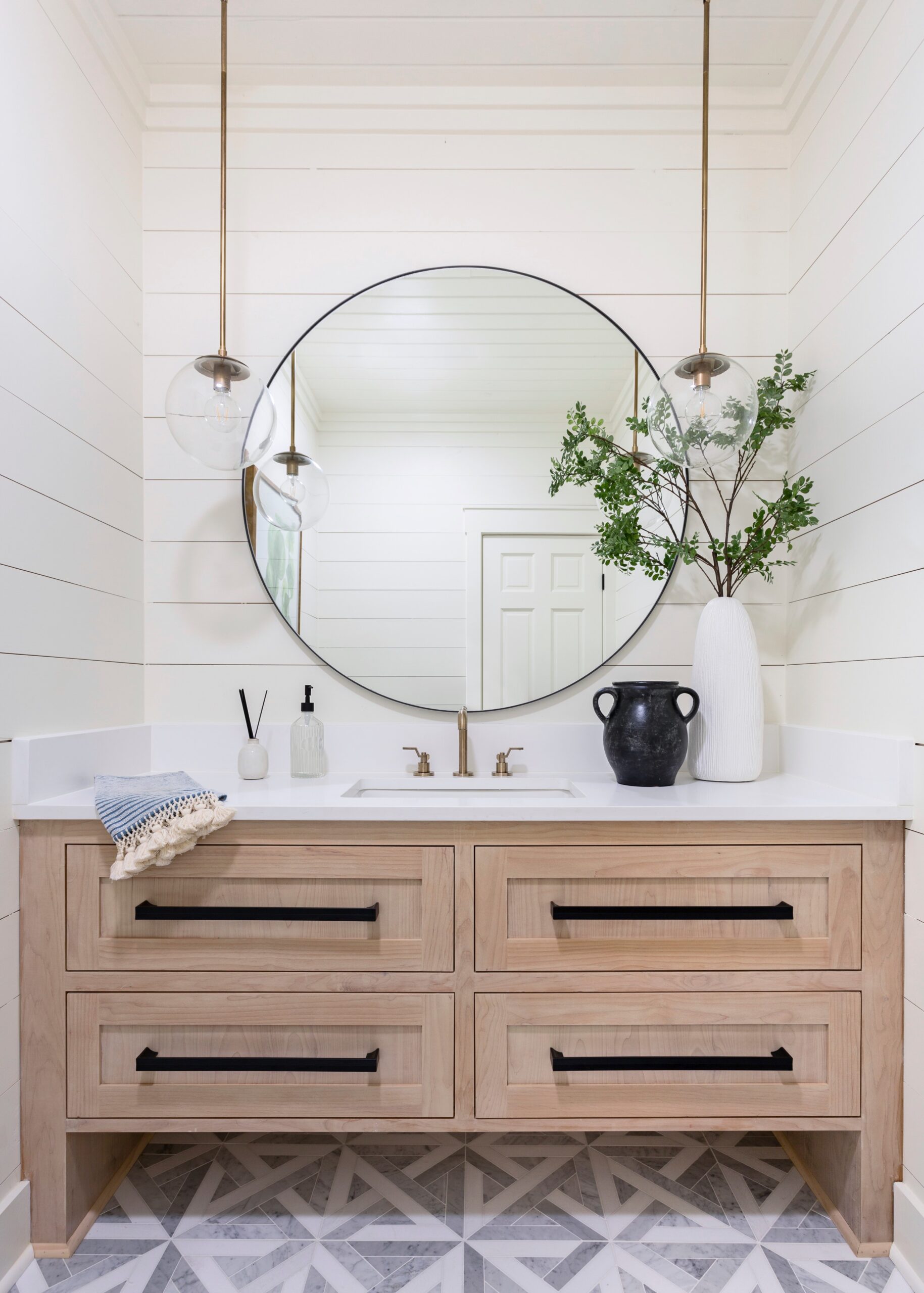 Modern farmhouse bathroom vanity with light wood cabinetry, black hardware, round mirror, and white shiplap walls.