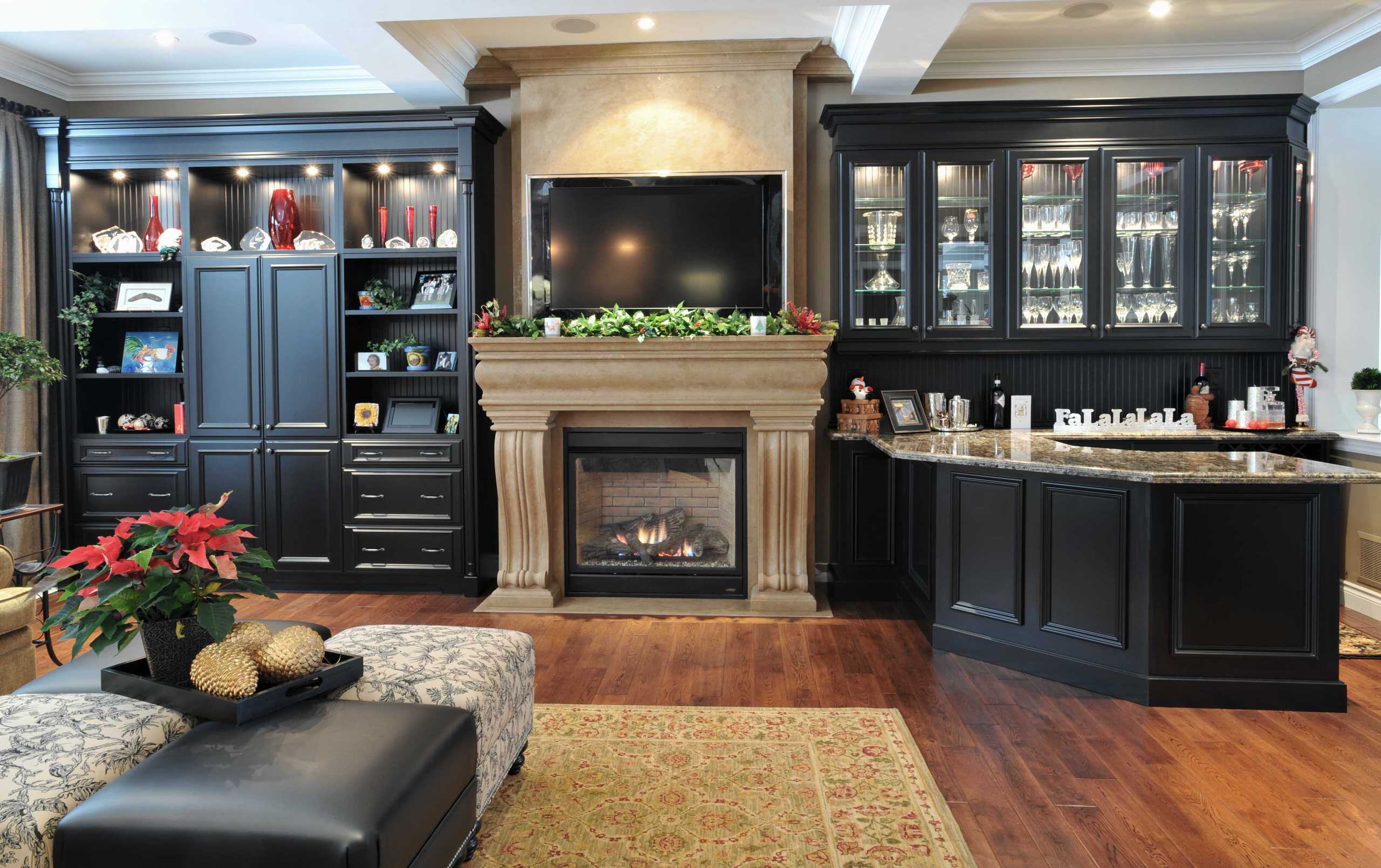A luxurious living room with a grand stone fireplace, a mounted flat-screen TV, custom black cabinetry with glass displays, a built-in bar, and elegant holiday decorations.
