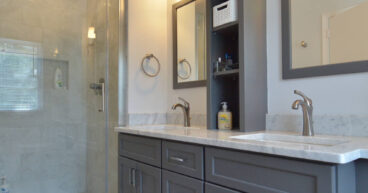 Modern bathroom with gray double vanity, marble countertop, built-in shelving, and a glass-enclosed shower with light beige tiles.
