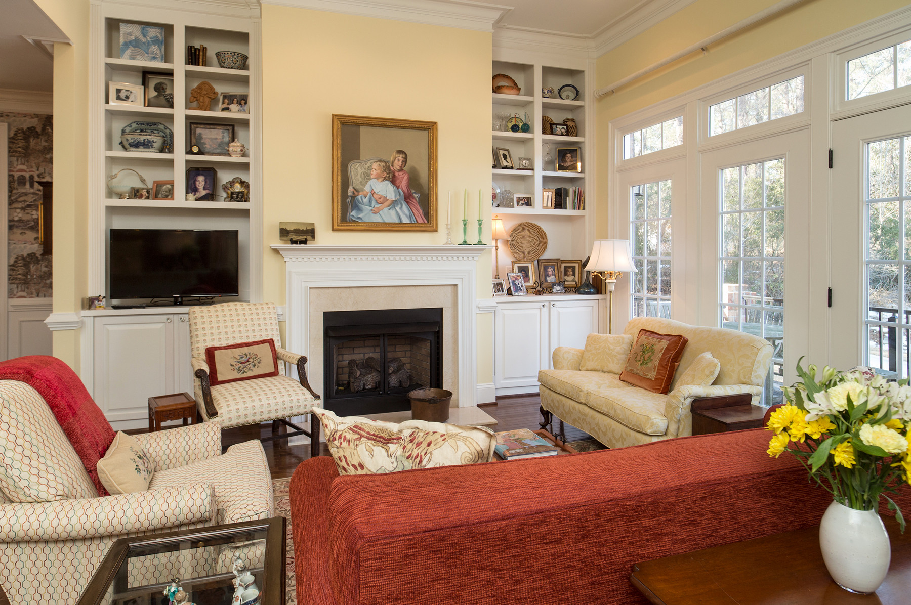 Traditional living room with a fireplace, built-in shelving, patterned armchairs, and large French doors letting in natural light.