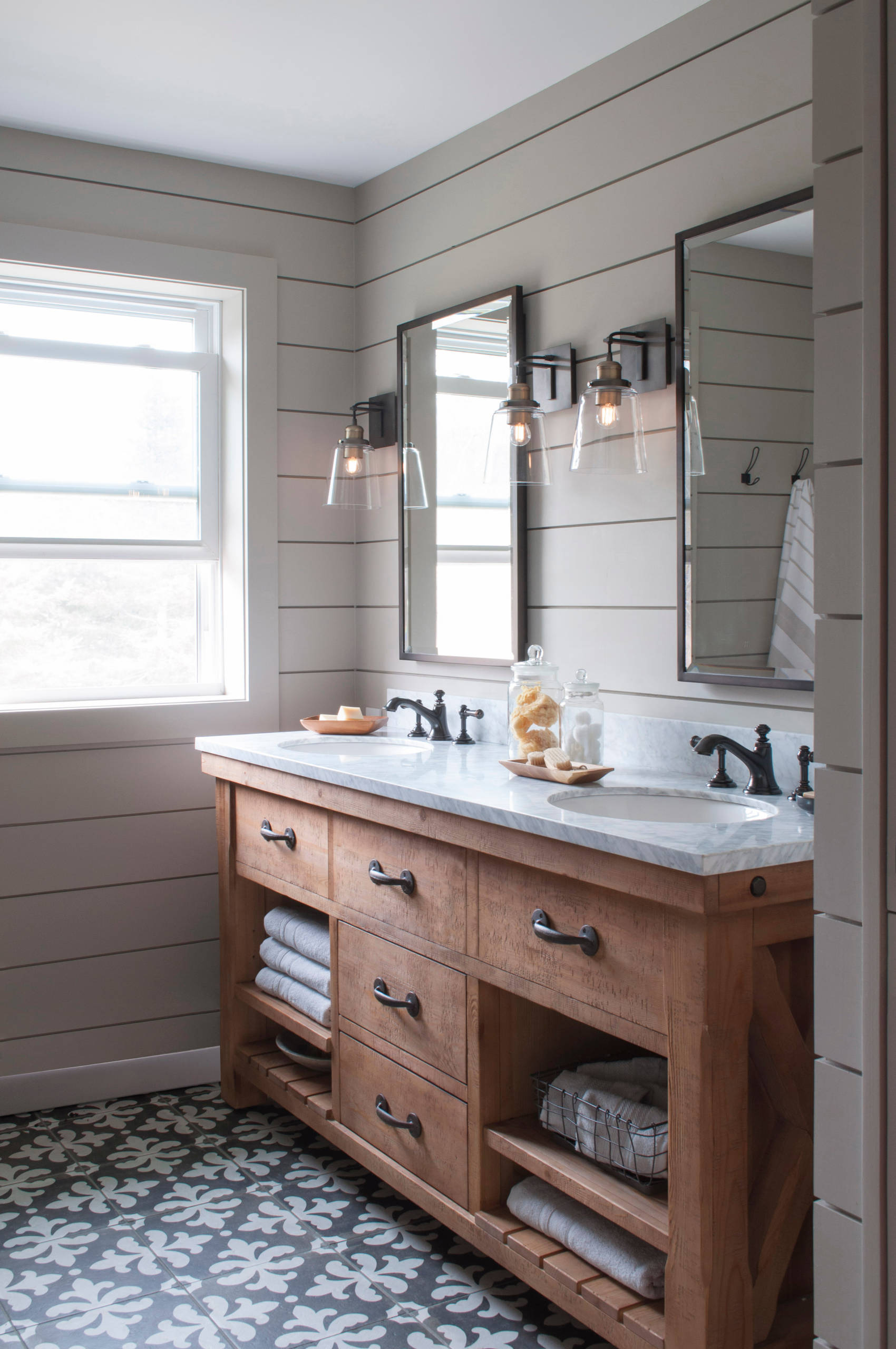 Rustic farmhouse bathroom with wooden vanity, double sinks, black fixtures, shiplap walls, and patterned tile floor.