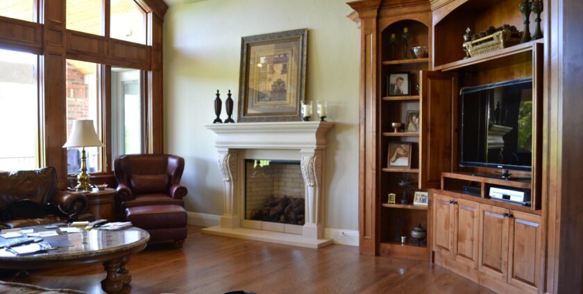 A traditional living room with a cream-colored ornate fireplace, large wood-framed windows, leather armchairs, a wooden entertainment unit with a TV, and hardwood flooring.