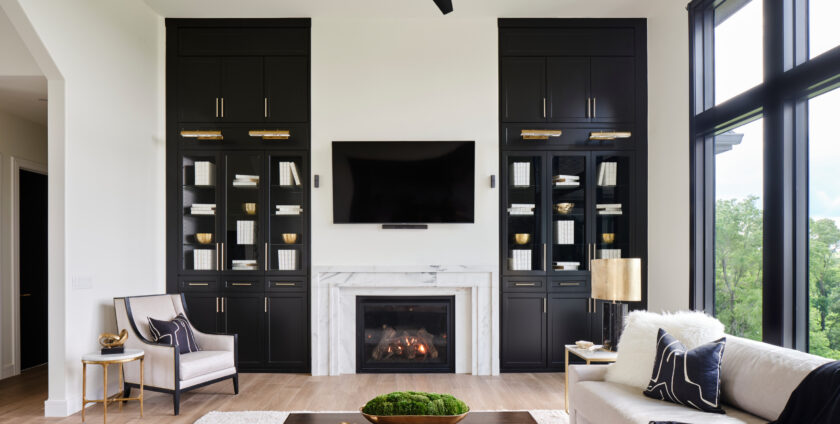 A modern living room with a white marble fireplace, black built-in cabinets, a mounted flat-screen TV, neutral sofas with accent pillows, and large windows providing natural light.