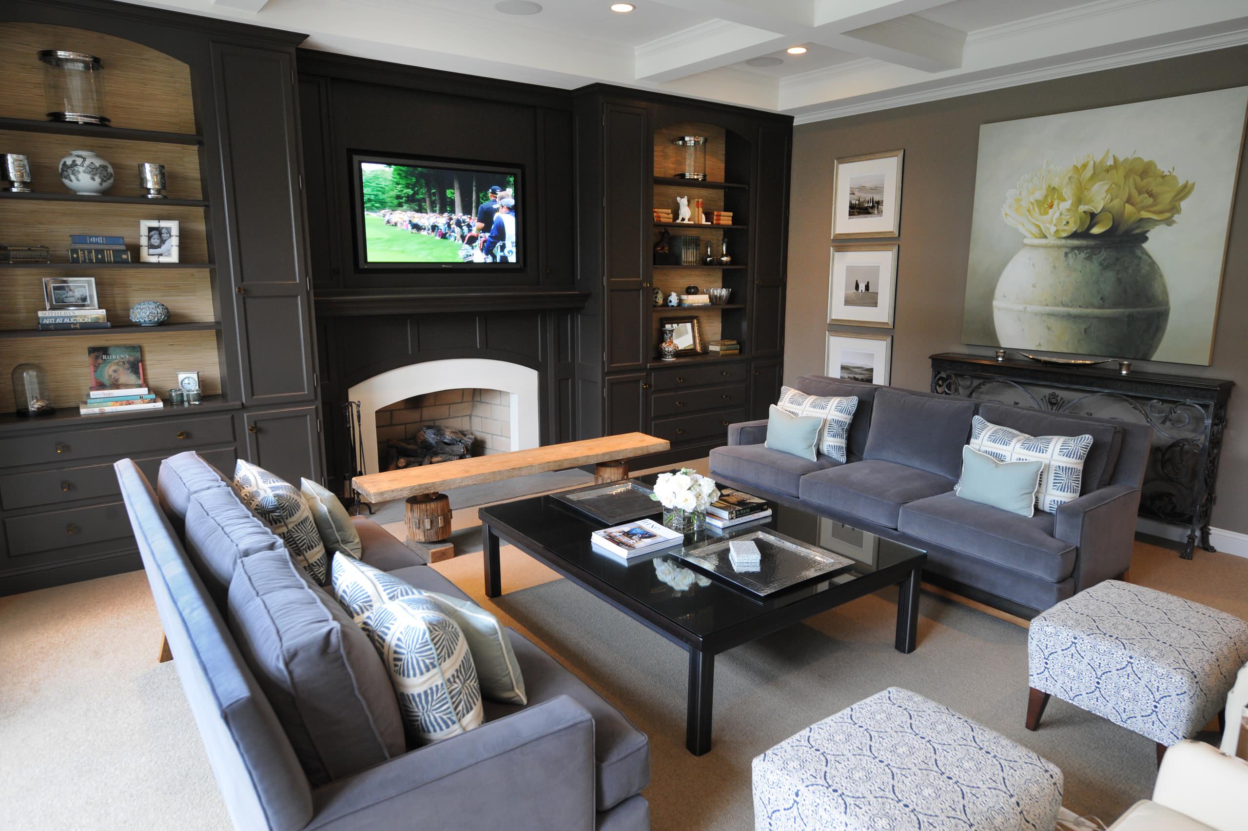 A modern living room with a white fireplace framed by dark built-in cabinets, gray sofas with patterned pillows, a glass coffee table, and a large floral painting on the wall.
