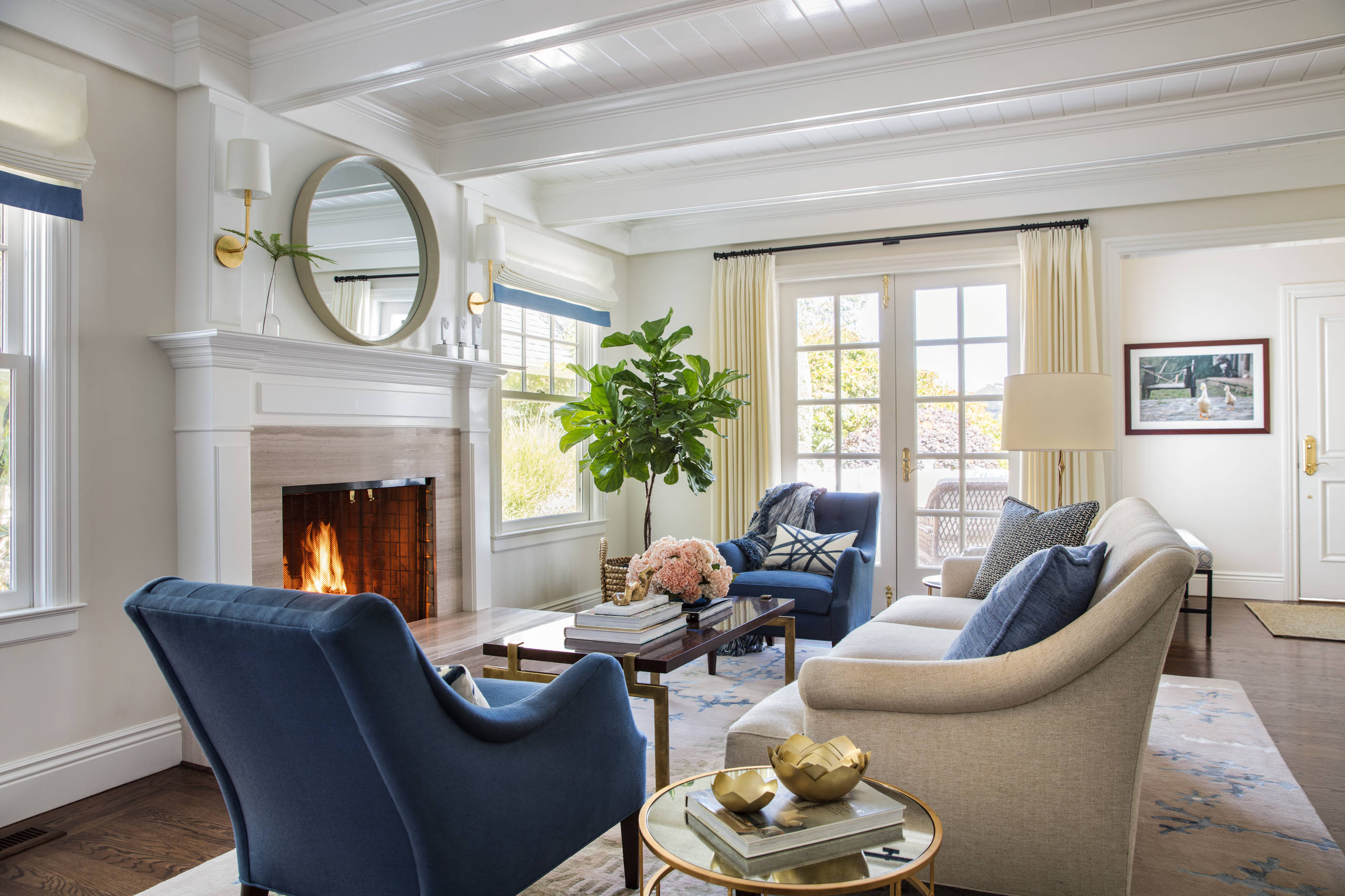 A bright living room with a white fireplace, a round mirror above the mantle, blue armchairs, a beige sofa, a coffee table with books and flowers, indoor plants, and large windows with cream curtains letting in natural light.