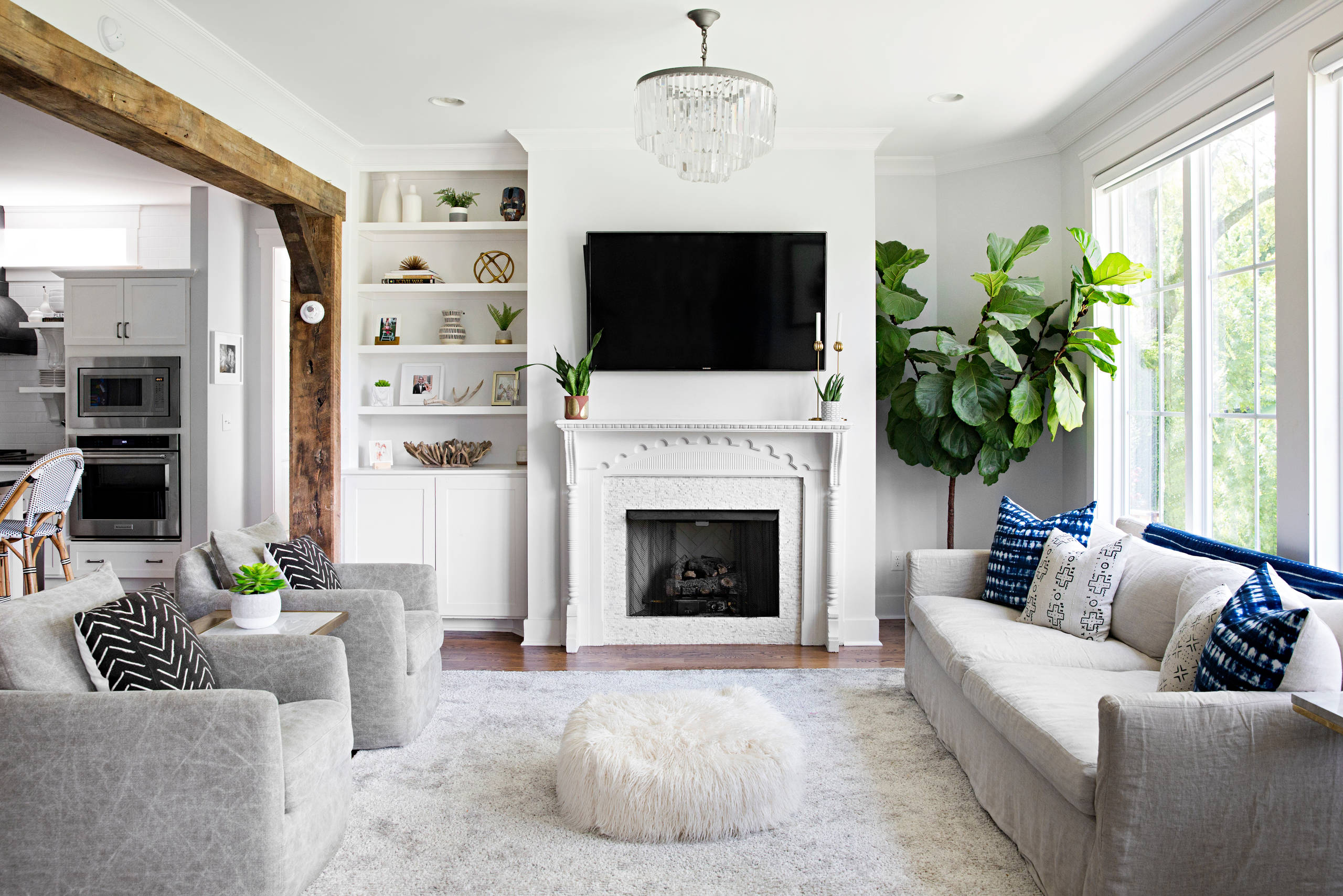 A bright living room with a white ornate fireplace, a mounted flat-screen TV, built-in shelves, gray armchairs, a white sofa with blue accent pillows, a fluffy ottoman, and large windows bringing in natural light.